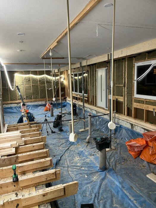 Renovering av hus med blottade bjälklag, väggar utan isolering och byggutrustning. Plastrullar täcker golvet, och det finns orange sopsäckar.