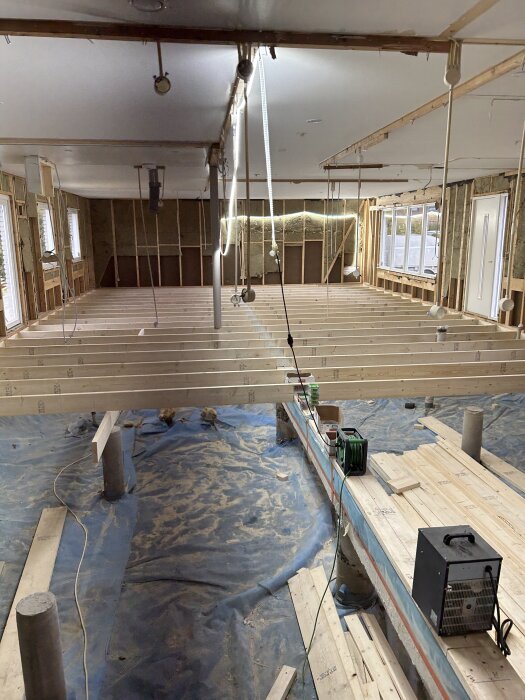 Renovering av byggnad inomhus, nyinstallation av bjälklag med synliga bjälkar och kablar. Trävirke, plastskydd på golvet och väggar delvis avlägsnade.