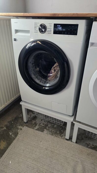 Vit frontmatad tvättmaskin på ett stativ, installerad bredvid en annan maskin, med kläder synliga genom dörren.