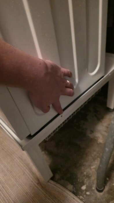 En hand håller på att öppna eller stänga en dörr till en vit frontmatad tvättmaskin, som står på ett betonggolv.