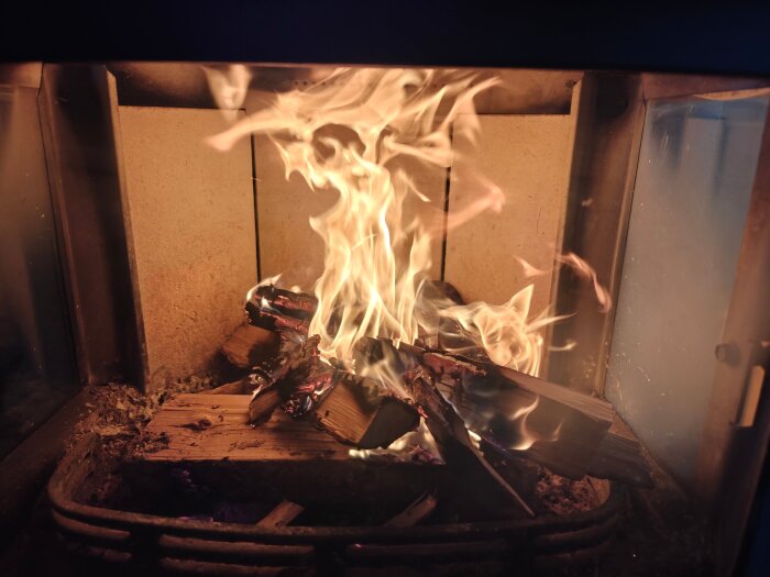 Brinnande ved i en kamin, med starka lågor och glödande träbitar. Kaminens inre är synligt med mörk ram runt glaset.