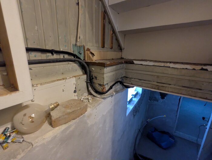 Slitet trävägg och kablar under en trappa, med en glödlampa och tegelsten på en betongavsats. Fönster med blått ljus syns i bakgrunden.