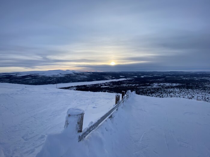 Snötäckt landskap i solnedgången, med berg i bakgrunden och ett staket i förgrunden.