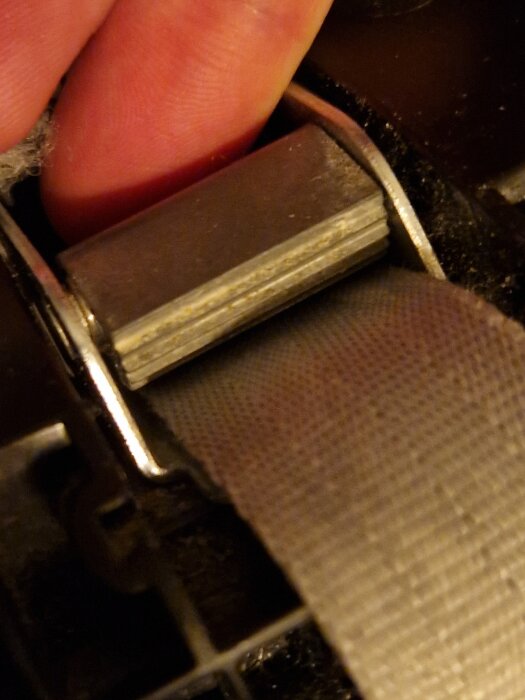 En hand justerar en metallspänne på ett band, troligen en del av en spännrem eller sele, mot en mörk bakgrund.
