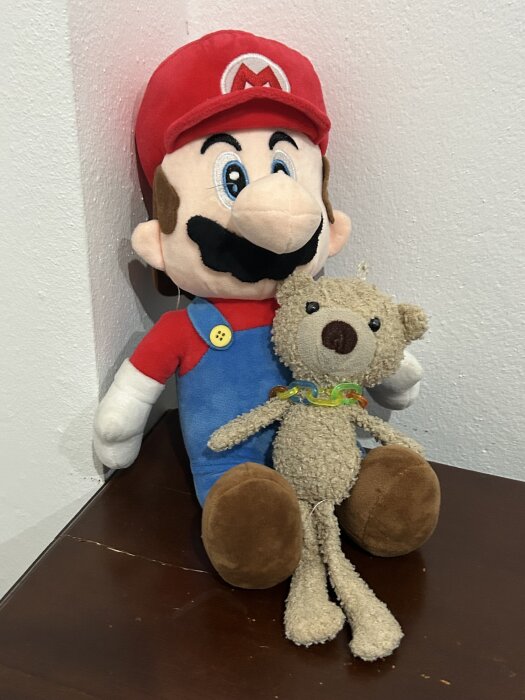 Leksaksfigur i röd mössa och blå hängselbyxor bredvid guldfärgad nallebjörn med flerfärgad halsband, sitter på ett mörkt träbord nära en vägg.
