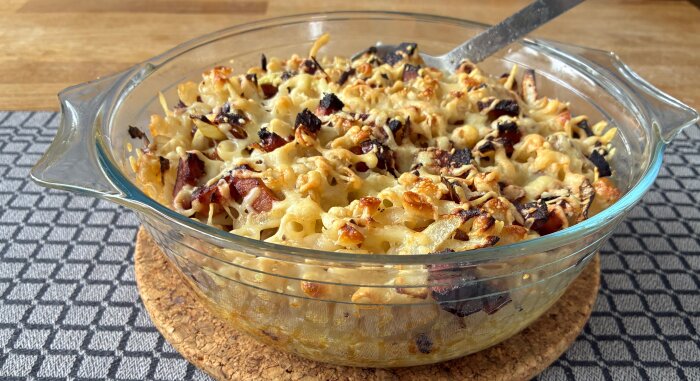 Gratinerad pastagratäng i glasform med smält ost, krispigt bacon och knaprig yta på en korkunderlägg.