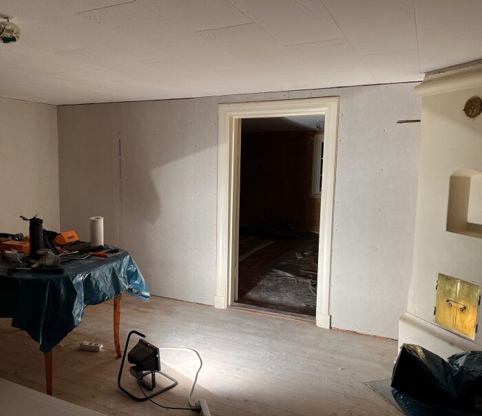 Renoverat rum med brädvägg täckt av ny gips, omålade foder runt dörröppning, arbetsbord med verktyg och belysning på golvet.