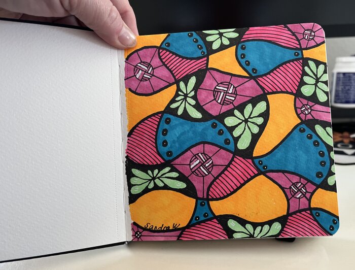 Färgglad teckning med abstrakta mönster i orange, blått, rosa och grönt med blommotiv. En hand håller i hörnet av boken.