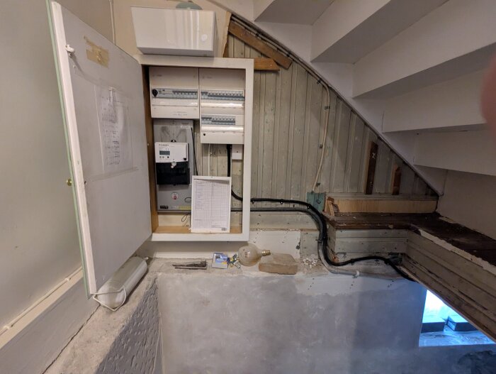Elcentral under trappa med öppen dörr som visar säkringar och ledningar; träpanel och böjar på rör synliga.