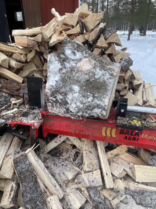 Vedkubbar och stora stockar staplade vid en vedklyv från Bala Agri, omgivna av snö.
