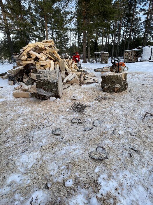 Vedhög av kapade stockbitar på snötäckt mark med sågspån och motorsåg i förgrunden, omgiven av skog.