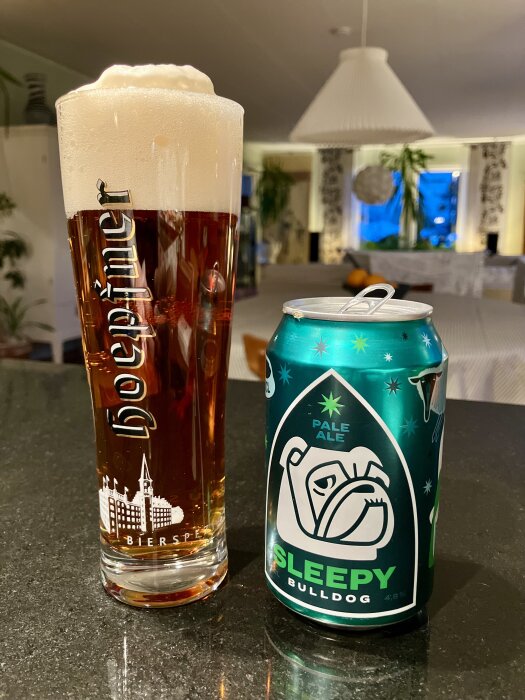 Ett glas med skummande öl bredvid en burk med "Sleepy Bulldog" pale ale på ett köksbord med bakgrund av en matsal.
