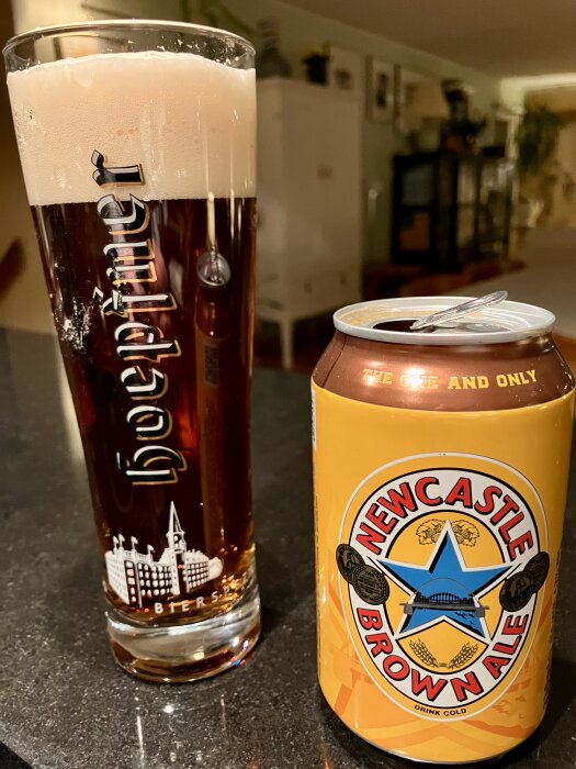 Ett glas mörk öl med tjockt skum bredvid en burk Newcastle Brown Ale på ett köksbord.