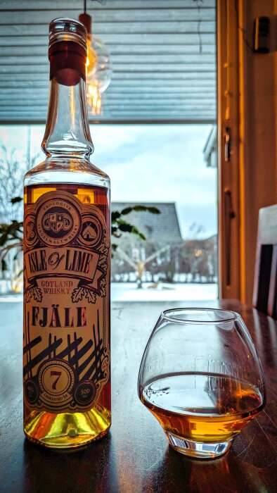 En flaska Isle of Lime whisky på ett bord med ett glas whisky bredvid, med ett snötäckt landskap utanför fönstret i bakgrunden.