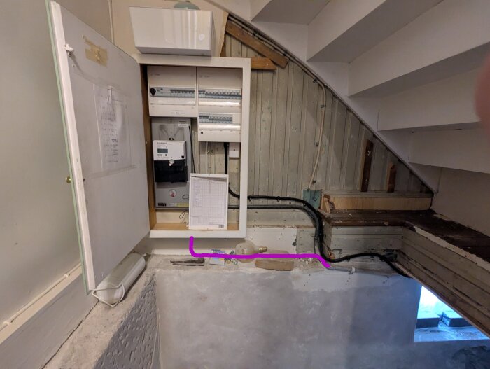 Elcentral under en trappa med kablar som går längs väggen. Öppna dörrar visar elkomponenter och en lista med installationsinformation.