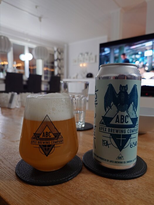 Ölglas och burk från Apex Brewing Company på ett bord, med en tv i bakgrunden.