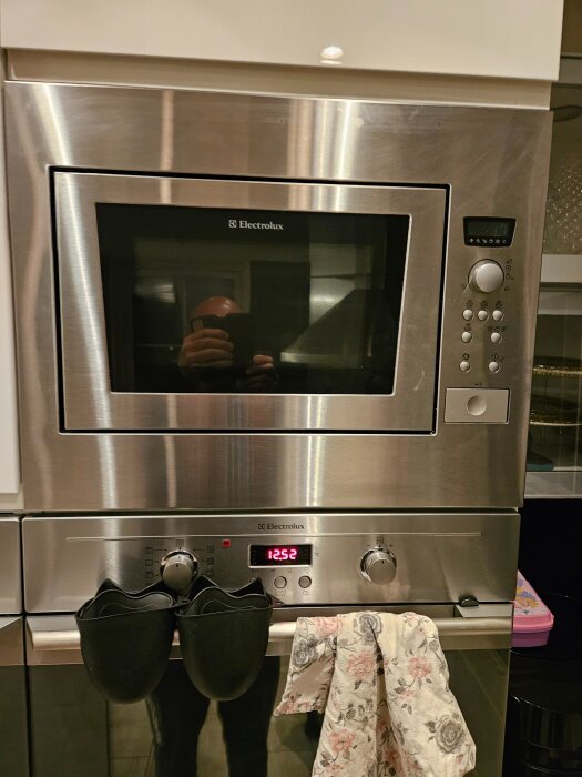 Inbyggd Electrolux-ugn med digital display och vred, två svarta muggar och en mönstrad kökshandduk hängande på handtaget.