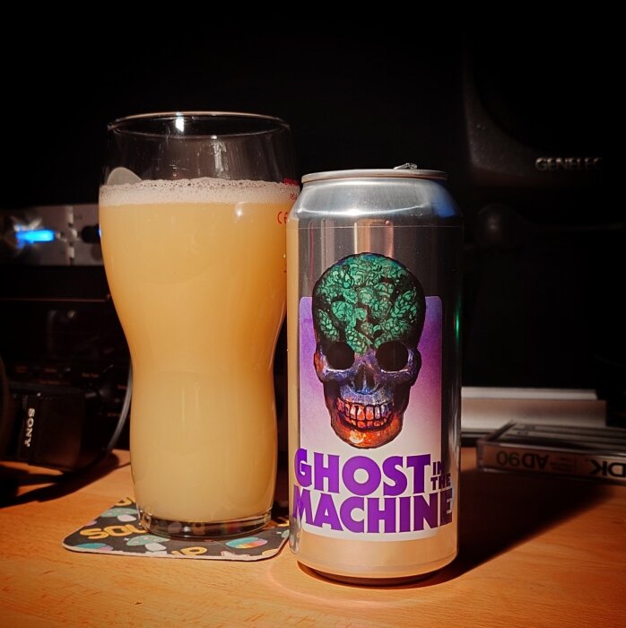 En ölburk med motiv av dödskalle och text "Ghost in the Machine" bredvid ett glas ljus öl på ett bord.