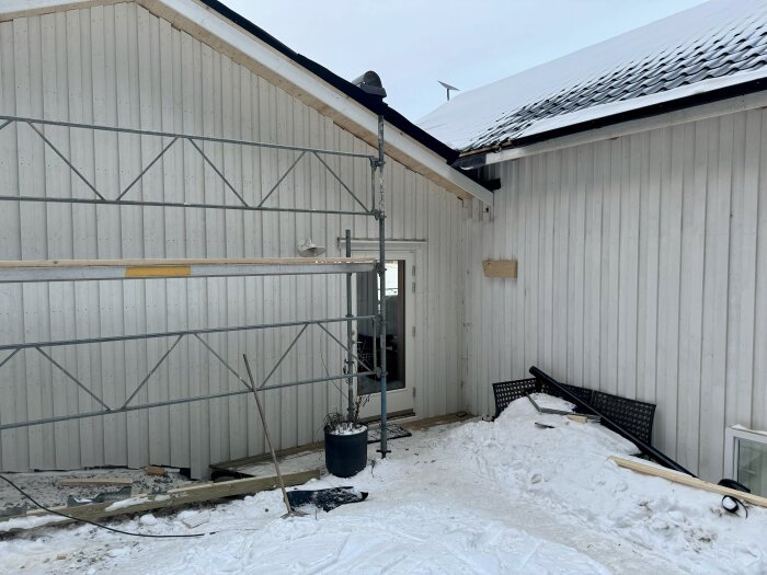 Vit träfasad med byggnadsställning, snö på marken och delar av tak syns. Saker intill väggen, redo för nästa byggfas.