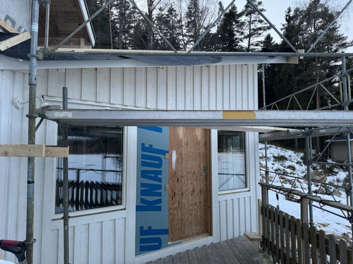 Byggnadsfasad med isolerat träpanel, byggställning runt om och en provisorisk dörr av plywood, vintermiljö i bakgrunden.