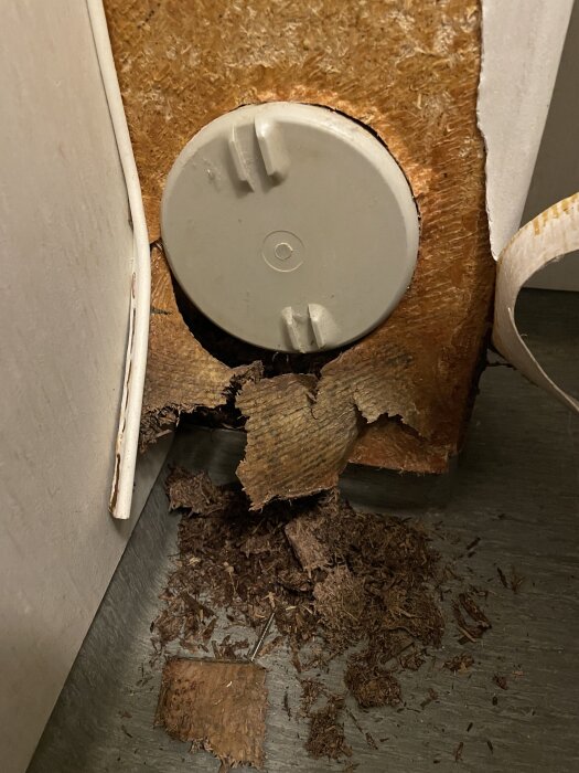 Rund renslucka på en vägg med skadad yta och träflisor på golvet, troligen från långvarigt läckage.