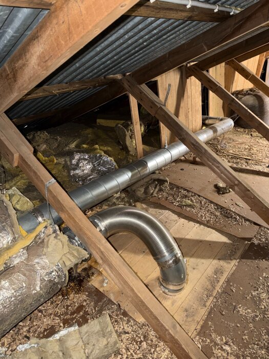 Ventilationsrör under ett tak i en isolerad vindsvåning, med träbjälkar och synlig metallbeklädnad, samt sågspån och isoleringsmaterial på golvet.