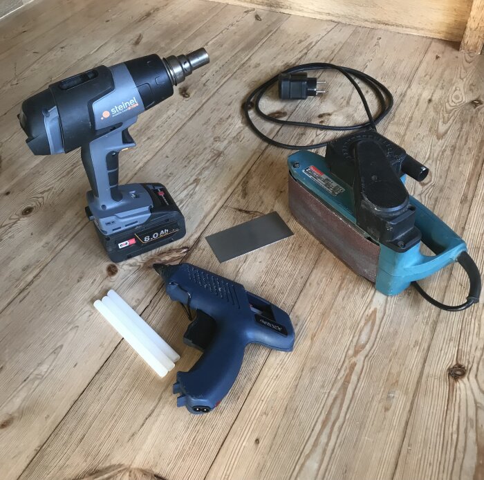 Verktyg för skidservice: varmluftspistol, bandslip, limpistol med polyetenstavar och en metallplatta, placerade på trägolv.