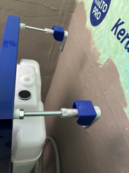 Närbild av en WC-fixtur med två för korta väggfästen monterade mot en brun vägg och en blå fixtur i ett smalt källarbadrum.
