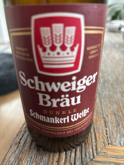 Flaska med etikett av Schweiger Bräu Dunkle Schmankerl Weiße öl, röd bakgrund med vit text och kronsymbol, står på ett träbord.