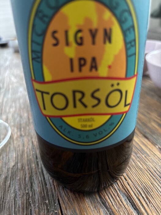 Öletikett på en flaska Sigyn IPA Torsöl med 5,6% alkohol, 500 ml volym, liggande på ett träbord.