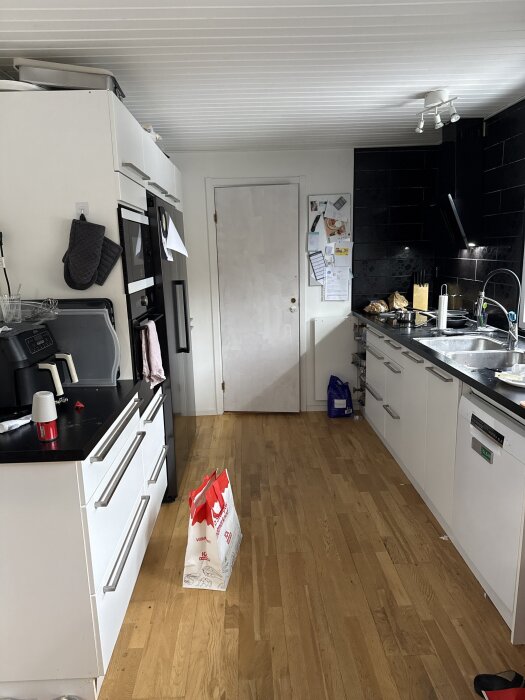 Modernt kök med vit inredning längs två väggar, trägolv, svarta kakelplattor och diverse köksredskap. En vit dörr i bakgrunden.