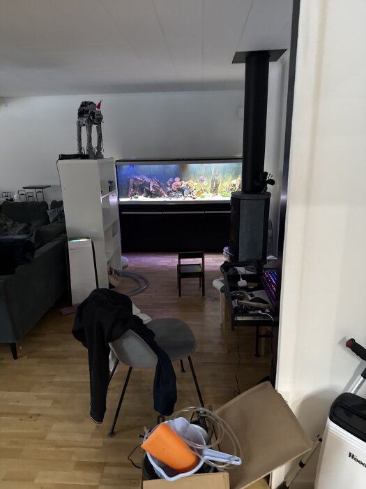 Rum med en akvarium på en bänk, stökiga ytor med stolar, kartonger och en kamin.