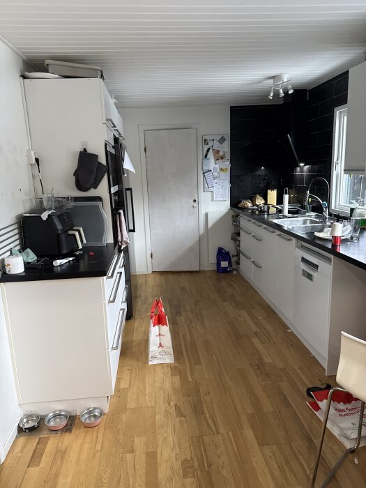 Kök med vita skåp och svart bänkskiva, träparkettgolv, och diskho till höger. Dörr och vägg prydd med lappar längst bak.
