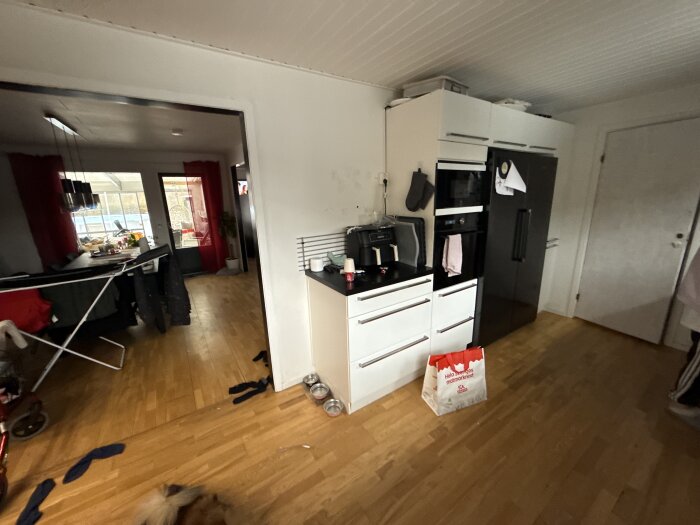 Kök med vita skåp, svart kyl och ugn. Tvål, handdukar och en papperspåse syns på golvet. Matplats i bakgrunden med rött draperi.