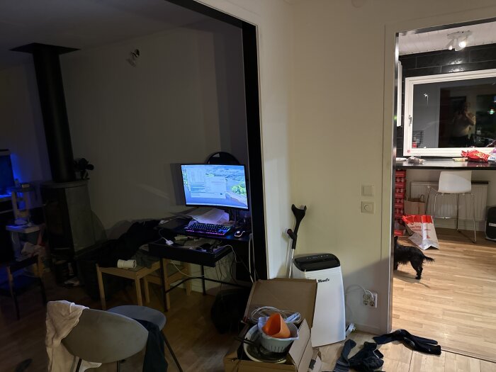 Rum med datorhörna, kamin, stökigt golv och ett kök i bakgrunden. En hund och diverse föremål syns.