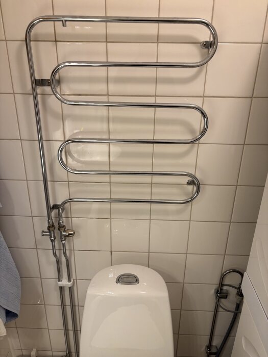 Kromad handdukstork monterad på en vägg i badrum, med rör och ventiler synliga ovanför en toalettstol.