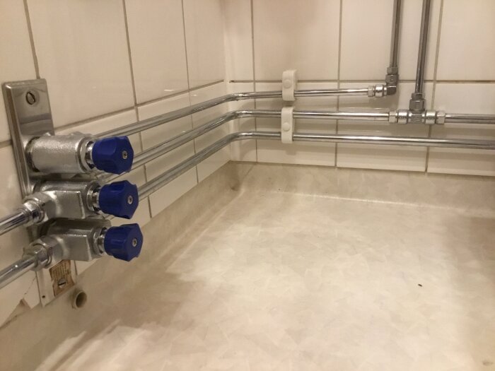 Tre vattenventiler med blåa vred monterade vid väggen med anslutna rör. Rör går åt vänster mot ett handfat.