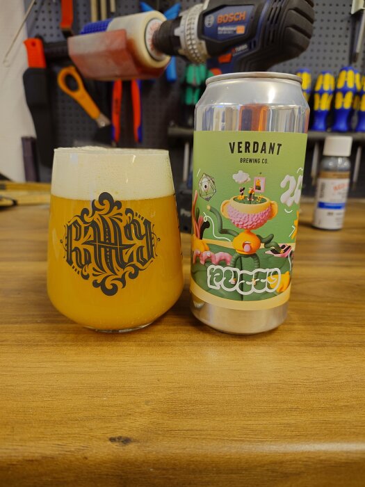 Ett glas med gyllene öl bredvid en burk från Verdant Brewing Co på en träbänk, med verktyg och en borrmaskin i bakgrunden.