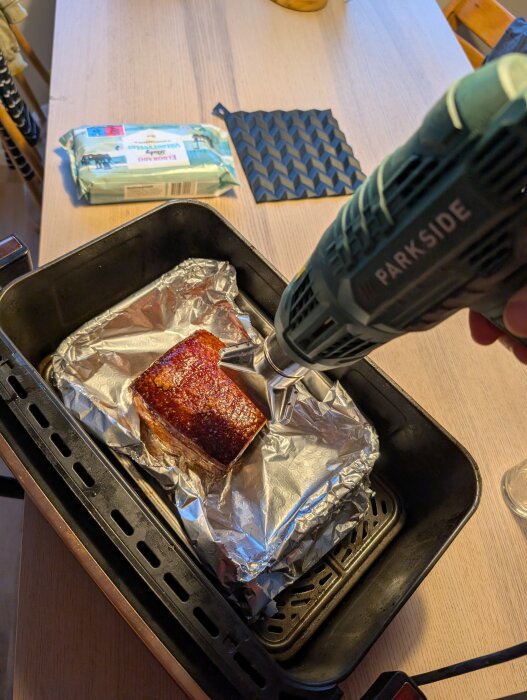 En fläsksida ligger i en ugnsform, täckt med folie, och bearbetas med ett elverktyg för att krispa upp svålen.