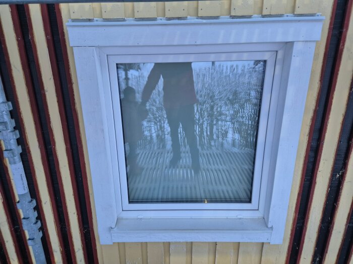 Fönster med vit fönsterkarm på randig yttervägg, reflektion av vuxen och barn synlig i glaset.