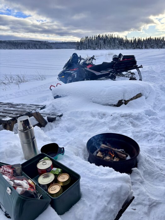 Snöskoter vid sjö, brasa i snön, matlåda med konserver och termos. Skog i bakgrunden, vinterlandskap.