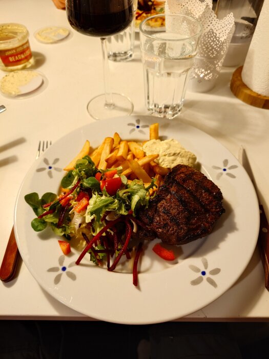Grillad rostas med pommes, sallad och bearnaisesås på tallrik. Vin- och vattenglas i bakgrunden.