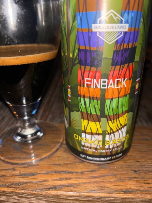 Flaska med fin färgsprakande etikett av "Omako Basoa Imperial Pastry Stout" bredvid ett glas med mörk stout på ett träbord.