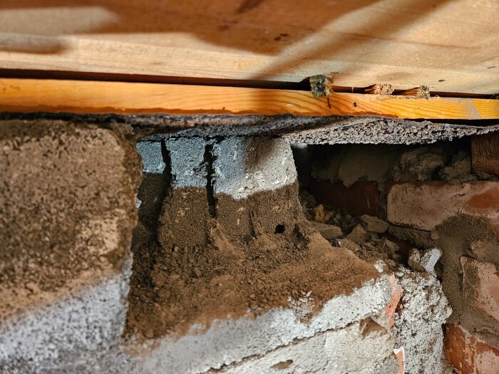 Närbild av en murad vägg med sprickor och lös cement under en träbjälke i ett byggprojekt.