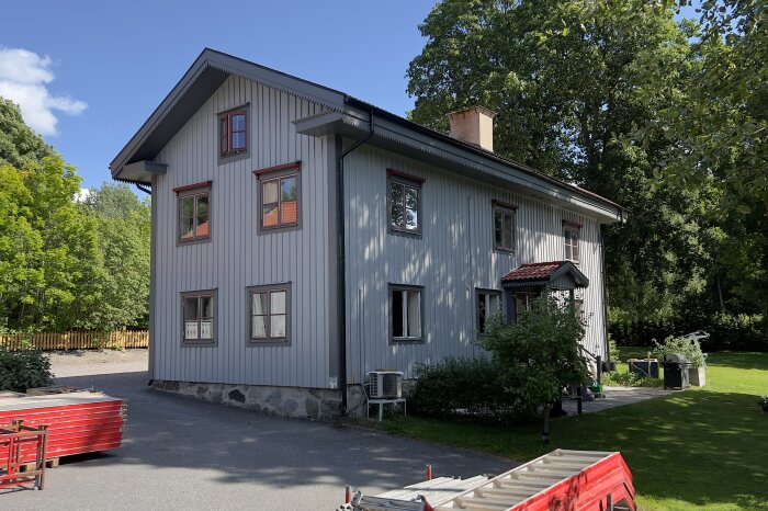Grått tvåvåningshus med rött tak, beläget i en grön trädgård. Soligt väder med blå himmel, stegar och arbetsverktyg synliga i förgrunden.