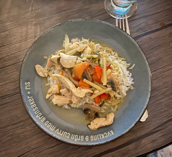 Kyckling i grön curry med grönsaker och ris på en tallrik, omgiven av bestick och ett glas vatten på ett träbord.