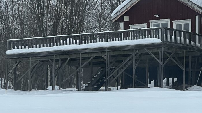 Altan framför ett rött hus, täckt av snö, med synliga träbjälkar under och träd i bakgrunden.