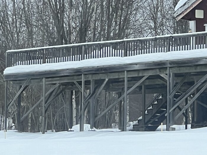 Träaltan täckt av snö, med trästolpar och trappa, omgiven av träd på vintrig bakgrund. Ser instabil ut.