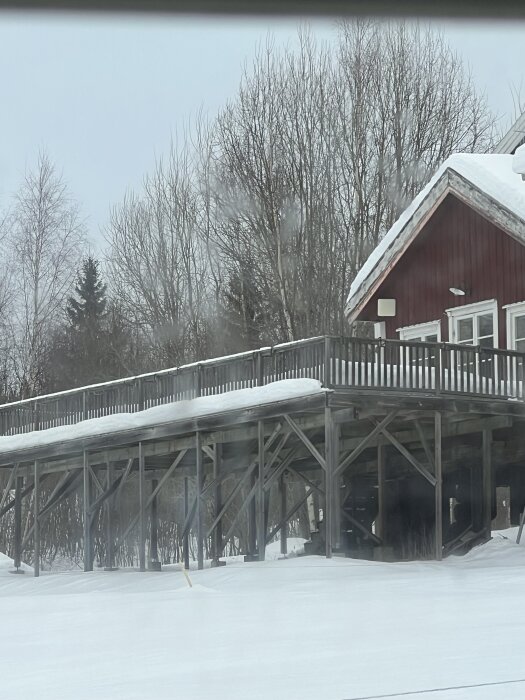 En stor altan med snöbelagd yta och trästöttor, belägen vid ett rött hus i ett snöigt landskap.