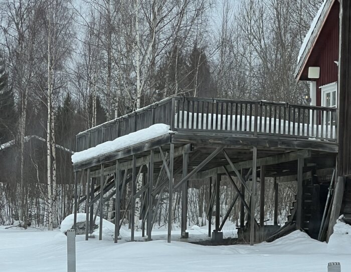 Altan med snö på räcket och pelare som stöder upphöjd konstruktion vid ett hus i vintrigt landskap.